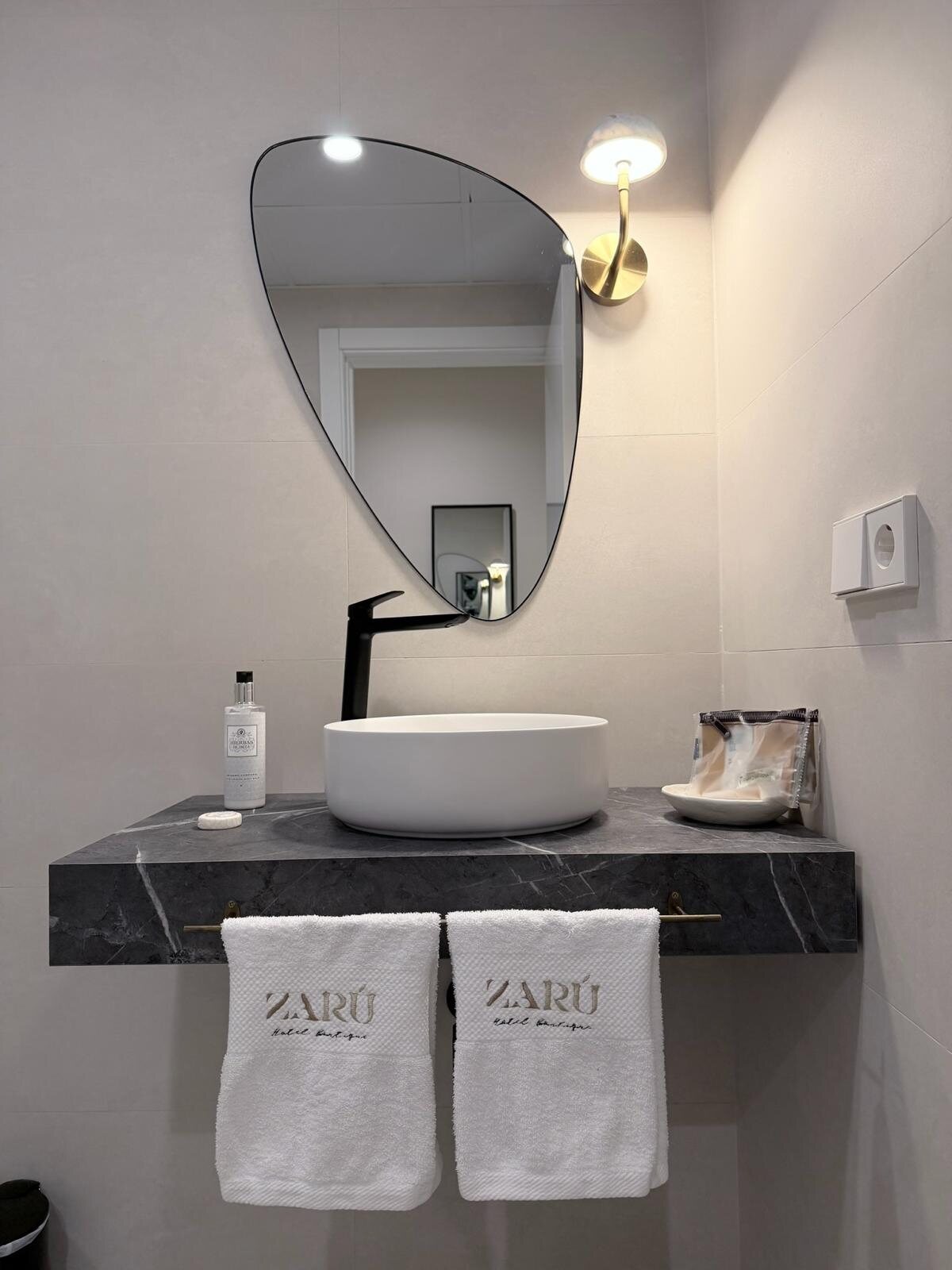 Baño del hotel Zarú con lavabo oscuro, espejo redondo y apliques